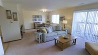 This single-floor residence offers a warm and inviting atmosphere, highlighted by an open-concept layout that seamlessly connects the living and dining areas. The living space is anchored by a comfortable striped sofa accented with decorative pillows, complemented by light blue end tables and a wooden coffee table that add a touch of casual charm. Soft, neutral-toned walls and plush wall-to-wall carpeting create a cozy and relaxed ambiance throughout. Natural light filters in through a large window fitted with vertical blinds, brightening the space and enhancing its airy feel. A ceiling-mounted light fixture provides additional illumination, while a classic table lamp adds a warm, ambient glow. Decorative elements, including a framed gallery wall arrangement and a tasteful floral arrangement, lend personality to the interior. The adjacent dining area accommodates a full dining set, making it well-suited for everyday meals or hosting guests. The open flow between the two areas makes the overall space feel expansive and functional, ideal for both relaxing and entertaining. The thoughtful layout and light color palette come together to create a comfortable, livable environment.