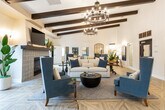 Located at 7750 Bolsa Ave in Midway City, California, this single-story community clubhouse offers a thoughtfully designed gathering space that blends Spanish-inspired architecture with contemporary interior styling. The main lounge is the centerpiece of the space, boasting soaring ceilings with prominent dark wood exposed beams and dramatic wrought-iron candle-style chandeliers that cast a warm, inviting glow. A decorative tiled fireplace anchors one wall beneath a mounted flat-screen television, while generous seating arrangements of plush sofas and wingback chairs in navy, cream, and gold create an atmosphere ideal for relaxed socializing. Intricate geometric parquet-patterned flooring runs throughout the main areas, adding a refined architectural detail underfoot. The fully equipped kitchen features deep slate-blue cabinetry paired with crisp white shaker-style lower cabinets, white quartz countertops, and a distinctive geometric wood-inlay floor. A large sliding window above the sink frames a direct view of the outdoor patio and surrounding landscaping, flooding the space with natural light. A separate private dining room provides an intimate setting with a substantial natural wood dining table, upholstered teal and neutral chairs, a bold dark accent wall, and an industrial-style pendant light. Multiple access points connect the interior seamlessly to the outdoor areas, enhancing the overall sense of openness and flow throughout this well-appointed community facility.