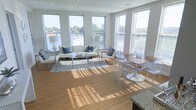 This single-floor space is designed to maximize natural light and an open, airy feel. Large, floor-to-ceiling style windows wrap around the main living area, flooding the space with abundant sunlight and offering pleasant outdoor views of the surrounding neighborhood. The warm hardwood flooring extends throughout, lending a cohesive and inviting aesthetic to the entire layout.

The living area is generously proportioned, accommodating a comfortable seating arrangement with a sleek, modern sofa and accent chairs centered around a glass coffee table. The open-concept design flows naturally into a dining area, where a wooden dining table is paired with contemporary acrylic ghost chairs, creating an elegant contrast between warm and modern elements.

Adjacent to the dining area, a glimpse of the kitchen reveals stone countertops, hinting at a well-appointed culinary space that integrates seamlessly with the rest of the layout. The neutral, light gray walls serve as a clean backdrop that complements the bright, sun-drenched atmosphere throughout.

Overall, the space exudes a modern, minimalist character with thoughtful design choices that balance comfort and style. The abundance of natural light, open floor plan, and cohesive material palette make this a bright and welcoming environment suited for both relaxed living and entertaining.