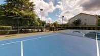 Located at 5715 Castlebridge Rd in Fredericksburg, Virginia, this single-story community amenity facility serves as a recreational hub for its surrounding residential neighborhood. The centerpiece of the property is a full-size, hard-surface tennis court finished in classic blue and green with crisp white court markings and a regulation net. Enclosed by a sturdy chain-link fence and equipped with overhead lighting, the court is designed for year-round play. Mature trees and well-maintained hedgerows border the court, providing natural shade and a pleasant, park-like atmosphere. Residential buildings are visible in the background, situating the facility comfortably within its neighborhood context. The interior of the facility is modest and functional, featuring tile flooring at the entrance accented by a wood-framed glass door that offers views toward the outdoor recreational area. Warm wood wainscoting runs along the interior walls, adding a touch of traditional character throughout the hallways and common spaces. Interior flooring transitions from tile at entry points to carpet in the common corridor areas. The facility includes clearly marked restrooms accessible from the main interior hallway, making it a convenient and well-equipped space for community members enjoying outdoor recreation.