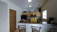 This two-story property includes a well-appointed kitchenette or hospitality area that serves as a welcoming hub for residents or visitors. The space features a wraparound bar-height counter with dark granite-style laminate surfaces, providing ample room for seating with black metal bar chairs positioned along the front. Above, light maple cabinetry lines the walls, offering generous storage, while the upper shelves display decorative lettering and a "Welcome to our home" sign, reinforcing the inviting atmosphere of the space.

The kitchenette is equipped with a full-size refrigerator and additional counter appliances, along with a secondary counter area near a window that allows natural light to filter in. Branded yellow cups and packaged snacks are displayed on the counter, suggesting this area functions as a communal amenity or leasing office refreshment station — consistent with the "The Row" branding visible in the room.

Flush-mount ceiling lights provide consistent overhead illumination throughout the space. The light tile flooring and neutral white walls keep the overall aesthetic clean and functional, while the warm wood cabinetry adds a residential touch to what is an organized, practical, and welcoming environment.