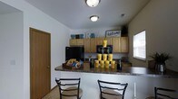 This two-story property includes a well-appointed kitchenette or hospitality area that serves as a welcoming hub for residents or visitors. The space features a wraparound bar-height counter with dark granite-style laminate surfaces, providing ample room for seating with black metal bar chairs positioned along the front. Above, light maple cabinetry lines the walls, offering generous storage, while the upper shelves display decorative lettering and a "Welcome to our home" sign, reinforcing the inviting atmosphere of the space.

The kitchenette is equipped with a full-size refrigerator and additional counter appliances, along with a secondary counter area near a window that allows natural light to filter in. Branded yellow cups and packaged snacks are displayed on the counter, suggesting this area functions as a communal amenity or leasing office refreshment station — consistent with the "The Row" branding visible in the room.

Flush-mount ceiling lights provide consistent overhead illumination throughout the space. The light tile flooring and neutral white walls keep the overall aesthetic clean and functional, while the warm wood cabinetry adds a residential touch to what is an organized, practical, and welcoming environment.