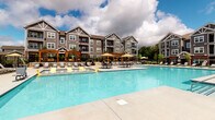 This outdoor amenity space centers around a generously sized resort-style swimming pool with crystal-clear blue water, designed to serve as a premier gathering and relaxation destination for residents. The pool deck is finished in smooth concrete and offers ample space for lounging, with rows of chaise lounge chairs positioned along the perimeter for sunbathing and leisure. Multiple umbrella-shaded seating areas are distributed throughout the deck, providing shaded retreats for outdoor dining and socializing. Elegant wooden pergola structures anchor one side of the deck, adding architectural character and a defined gathering zone with additional table and chair seating beneath. The pool area is enclosed by a sleek metal fence, ensuring a secure and private environment. The exterior building facade that surrounds the space features a craftsman-inspired architectural style, characterized by gray horizontal siding, white trim detailing, gabled rooflines, and brick accents at the base — creating a cohesive and attractive visual backdrop. Large windows overlook the pool from adjacent indoor spaces, and decorative planters with colorful seasonal flowers add lively touches throughout. The overall ambiance is open, bright, and inviting, well-suited for outdoor recreation and community engagement.