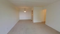 This single-floor space presents a straightforward and functional layout centered around an open living area. The room is finished with wall-to-wall neutral-toned carpeting that extends throughout, contributing to a soft and cohesive feel underfoot. The walls are painted in a clean, bright white, helping to reflect light and maintain an airy atmosphere throughout the interior.

A pendant light fixture hangs from the ceiling, positioned toward the dining or central area of the main room, providing focused overhead illumination. The ceiling is standard height and features a subtle recessed border detail, adding a modest architectural element to the otherwise simple design.

The layout opens toward additional spaces through a combination of doorways and open archways. One doorway leads to what appears to be a kitchen area, visible in the background with cabinetry and appliances hinting at its function. An arched opening on the right side of the main room connects to another area of the unit, suggesting a versatile, open-flow floor plan.

Overall, the space offers a clean, neutral canvas that is well-suited for a variety of furnishing styles, with its open configuration supporting easy movement between the main living areas.