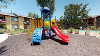 This residential community's outdoor grounds offer a thoughtfully designed environment with a variety of amenities suited for active, outdoor living. At the heart of the space are two colorful playground structures, each equipped with a bold red spiral slide, climbing elements, and multi-level platforms set over a cushioned gravel surface — ideal for children of all ages. The playgrounds are framed by mature shade trees that provide natural canopy and relief from the sun.

Adjacent to the play areas, a shimmering swimming pool is enclosed by a decorative wrought iron fence accented with circular detailing, with palm trees and tropical plantings adding a resort-like feel to the poolside setting. A tall wooden privacy fence defines the perimeter of the property, offering a sense of seclusion from surrounding areas.

The central courtyard features freshly mulched planting beds with low-maintenance shrubs and ornamental trees, connected by clean paved pathways that wind between the residential buildings. The warm-toned stucco exteriors of the surrounding multi-story buildings, complete with exterior staircases and private balconies, contribute to the cohesive architectural character of the community. The overall ambiance is bright, open, and family-friendly.