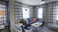 This single-floor space showcases a thoughtfully designed living area that blends contemporary style with comfortable functionality. The living room serves as the focal point, featuring a large dark gray tufted sectional sofa accented with a mix of colorful throw pillows, including coral and blue tones, that add warmth and personality to the space. A matching patterned area rug ties the room together and complements the bold geometric curtains that frame multiple windows along the walls. Natural light flows generously into the room through the large windows, which are dressed with stylish lattice-patterned drapes in a neutral gray and white palette. A sleek, industrial-style coffee table with a metal frame and wood top anchors the seating arrangement, while a modern arc floor lamp provides additional ambient lighting. A ceiling fan overhead ensures year-round comfort. The overall ambiance is modern yet inviting, with a cohesive design palette of grays, whites, and neutral tones punctuated by vibrant accent colors. The layout makes efficient use of space, creating a living environment that feels both open and cozy.