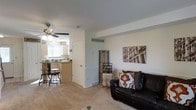 This two-floor residence offers a practical and comfortable layout with distinct living and kitchen areas visible from a single vantage point. The living area features wall-to-wall carpeting, neutral painted walls with a decorative chair-rail detail, and a leather sofa accented with decorative pillows. Artwork adorns the walls, adding character to the space. A ceiling fan with integrated lighting overhead provides both ventilation and ambient illumination. Adjacent to the living area, the kitchen is separated by a partial wall and doorway, featuring light wood cabinetry, a countertop bar with bar stools, and a window that allows natural light to filter into the space. A tall decorative cabinet adds a vintage touch near the room's dividing wall. The floor plan encourages easy flow between the living and kitchen areas while maintaining defined zones. A wall-mounted HVAC vent is visible, indicating central climate control. The overall ambiance is casual and livable, with a neutral palette that provides a versatile backdrop suitable for a variety of interior styles.