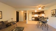 This single-floor residence offers an open-concept layout that seamlessly connects the living area to the kitchen and dining space. The living room features carpeted flooring and is furnished with comfortable seating, providing a relaxed and welcoming atmosphere. A ceiling fan with an integrated light fixture is centrally positioned overhead, ensuring both comfort and illumination throughout the shared space.

The kitchen is equipped with dark-toned cabinetry, a full-size refrigerator, a dishwasher, and a range, offering ample storage and functionality. A bar-height dining table with accompanying stools is situated adjacent to the kitchen, creating a convenient casual dining area. The kitchen floor transitions to a lighter tile or linoleum surface, clearly delineating it from the carpeted living area.

A structural column serves as a subtle visual divider between the living and kitchen zones without disrupting the open feel of the space. A hallway accessible from the main living area leads to additional rooms, including what appears to be further living quarters. A solid front entry door with a dark finish anchors the entryway, contributing to the overall sense of privacy and security. The warm, ambient lighting throughout gives the space an inviting and cozy character.