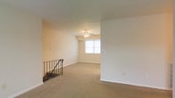 This single-floor residential space presents a clean, functional layout designed for comfortable everyday living. The main living area is finished with wall-to-wall carpeting in a warm neutral tone, contributing to a soft and inviting atmosphere throughout. Smooth white walls and a white ceiling create a bright, airy feel that enhances the sense of openness in the room. A ceiling fan with an integrated light fixture is centrally positioned, providing both ventilation and ambient lighting. A window along the far wall allows natural light to filter into the space, further brightening the interior. A staircase with a classic wrought iron railing is visible, adding a subtle decorative touch and indicating vertical access within the building. The neutral color palette and straightforward design of the space offer a versatile backdrop that can accommodate a variety of furnishing styles and personal preferences. Overall, the space conveys a practical and comfortable residential environment suited for a range of occupants.