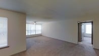 This single-floor space features a generously sized open living area finished with wall-to-wall carpet in a neutral beige tone, lending a clean and understated aesthetic throughout. The room is illuminated by natural light streaming through a large window, which frames views of mature trees outside, creating a connection to the outdoors. A ceiling fan is centrally positioned overhead, providing both air circulation and a modest focal point to the otherwise minimalist ceiling. The walls are painted in a clean white, contributing to a bright and airy atmosphere. The layout is straightforward and open, offering flexible potential for furniture arrangement. Subtle baseboard trim runs along the perimeter of the room, adding a refined finishing touch. The overall ambiance is calm and neutral, making it a versatile canvas well-suited to a variety of interior design preferences.