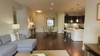 This single-floor residence showcases an open-concept layout that effortlessly connects the living, dining, and kitchen areas into one cohesive and functional space. Rich, dark hardwood flooring extends throughout, anchoring the design with warmth and continuity.

The living area offers a comfortable, understated setting, while the dining space accommodates a glass-top table surrounded by upholstered chairs, positioned beneath recessed lighting that ensures a well-lit atmosphere. A window at the rear of the dining area introduces natural light into the heart of the space.

The kitchen is equipped with dark cabinetry contrasted against light countertops and modern stainless steel appliances, including a built-in microwave. A peninsula with bar-stool seating serves as both a functional workspace and a casual gathering spot. Pendant lighting above the peninsula adds a refined touch to the kitchen's overall aesthetic.

Neutral-toned walls, recessed lighting throughout, and a thoughtful layout contribute to a clean, modern ambiance. The open floor plan maximizes the sense of spaciousness, making the space feel both inviting and practical for everyday living.