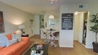 This single-floor space showcases an open-concept layout that seamlessly connects the living, dining, and kitchen areas into one cohesive environment. The living area is anchored by a warm-toned sofa and a dark coffee table, creating a relaxed and inviting atmosphere. Plush carpeting extends throughout, adding warmth and comfort underfoot.

A bistro-style dining nook sits centrally between the living and kitchen areas, featuring a sleek round high-top table paired with modern curved bar stools. A pendant light hangs overhead, drawing attention to this intimate dining space and adding a touch of charm.

The kitchen is partially open to the main living area via a pass-through counter, creating a sense of connectivity while maintaining a defined cooking space. White cabinetry and bright lighting keep the kitchen feeling clean and functional.

The textured ceiling adds architectural character throughout the space, while neutral wall tones in soft greens and whites provide a calm backdrop. Decorative elements, including wall art and curated accessories, add personality and a lived-in warmth to the overall ambiance.