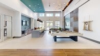 The Solaris Clubhouse in Kissimmee, Florida is a thoughtfully designed community facility that blends contemporary aesthetics with functional amenity spaces. The grand entrance lobby immediately impresses with soaring vaulted ceilings accented by teal coffers, large globe pendant chandeliers, and a bold chevron-patterned tile floor. Abundant natural light filters through clerestory windows and floor-to-ceiling glass doors, creating a bright and welcoming atmosphere. A reception desk and comfortable seating arrangements make the lobby an ideal gathering and welcome area. Beyond the entry, the expansive amenity lounge opens up with equally dramatic double-height ceilings highlighted by teal-painted recessed sections and stylish pendant lighting. This versatile space features a full-size billiards table, plush lounge seating anchored by area rugs, a sleek bar-height island with modern counter stools, and a wet bar with stainless steel appliances. Decorative lattice-panel windows and large exterior-facing glass flood the room with natural light and views of the surrounding community. Two well-proportioned hallways connect the main spaces, finished with clean white walls, large-format tile flooring, and tasteful artwork. Throughout the clubhouse, warm wood accents, curated décor, and cohesive neutral tones contribute to an upscale yet inviting ambiance.