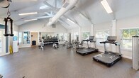 The fitness center at 800 Alexander Rd in Cayce, South Carolina is a single-floor exercise facility designed to support a comprehensive workout experience. The space is housed within an architecturally distinctive building featuring soaring vaulted ceilings with exposed white structural beams, which lend an open, airy feel throughout. Durable speckled rubber flooring covers the entire exercise area, providing both safety and comfort underfoot.

The facility is well-lit by a combination of recessed and overhead fluorescent lighting, supplemented by abundant natural light streaming through multiple windows fitted with blinds. Large glass doors open to an outdoor area, offering views of the surrounding greenery and pool area.

The exercise room is comprehensively equipped with a row of treadmills and elliptical machines positioned along the windowed walls, allowing users to enjoy outdoor views during cardio sessions. A dedicated strength training zone features cable and resistance machines, while a free weights area includes a full rack of dumbbells paired with adjustable workout benches. Large wall-mounted mirrors enhance the sense of space and assist with form during workouts.

Additional amenities include a water cooler station and security cameras. The neutral color palette of white and soft gray walls, accented by teal feature walls, creates a clean and motivating environment throughout the fitness center.