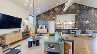 Located at 7900 SE Luther Road in Portland, Oregon, this 1,070 square foot single-story space offers a thoughtfully designed open-plan interior that blends modern aesthetics with warm, inviting character. The space is anchored by dramatic vaulted ceilings that soar overhead, accentuated by exposed structural beams and a mix of pendant and track lighting, including a statement multi-arm chandelier that adds visual interest. Light hardwood flooring flows throughout, lending warmth and continuity across the entire floor plan. A standout feature is the bold, floor-to-ceiling botanical wallcovering in deep navy, gold, and teal tones, which serves as a striking focal point and artistic centerpiece. The open living and lounge area is generously proportioned, accommodating multiple seating arrangements, including a green leather sofa, accent chairs, and coffee tables. Large black-framed windows invite abundant natural light and frame lush green tree views from multiple vantage points. A built-in media console with a wall-mounted flat-screen television anchors one end of the space, while a dedicated dining or café area with round tables and leather chairs provides a functional gathering zone. A kitchenette or service counter along the wall adds practical utility. Large glass doors open to an adjacent space, extending the sense of openness and connectivity.
