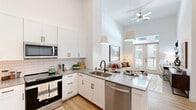 Located at 6801 Burnet Rd in Austin's vibrant 78757 neighborhood, this one-bedroom, one-bathroom residence offers a thoughtfully designed open-concept layout with impressive architectural details. Soaring ceilings in the kitchen and living area create an expansive, airy atmosphere, accentuated by large pendant lights and a ceiling fan. The kitchen is well-appointed with white shaker cabinetry, a white subway tile backsplash, stainless steel appliances including a refrigerator, range, microwave, and dishwasher, and a quartz-topped peninsula with bar seating — ideal for casual dining or entertaining. The living area flows seamlessly from the kitchen and opens to the outdoors through full-height French doors, flooding the space with natural light. Light wood-style flooring runs throughout the main areas, adding warmth and continuity. The bedroom offers a cozy carpeted retreat with a natural-light-filled window, while an en-suite bathroom features a sleek vanity with quartz countertops, a large black-framed mirror, and clean white tile accents. A dedicated walk-in closet with built-in shelving and hanging rods provides generous storage. The overall aesthetic is clean, contemporary, and bright throughout.