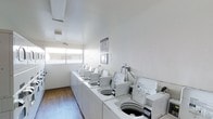 This single-floor communal laundry facility offers a clean and efficient environment designed for practical, everyday use. The room features a row of top-loading washing machines neatly aligned along one wall, providing ample capacity to serve multiple users. The machines are uniformly white, contributing to the room's bright, orderly appearance. Natural light streams in through a window on the adjacent wall, enhancing the open and airy feel of the space. The flooring consists of hardwood-style planks that add a touch of warmth to the otherwise utilitarian setting. The walls are painted a crisp white, maintaining a clean and hygienic atmosphere throughout. A posted notice on the wall provides users with guidelines and operating hours, ensuring the facility is used responsibly. The ceiling is smooth and unadorned, keeping the focus on the room's functional elements. Overall, this laundry facility is thoughtfully laid out to maximize usability, with sufficient space to move comfortably between machines. Its well-maintained condition and bright ambiance make it a convenient and welcoming amenity.