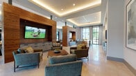 This elegantly designed single-floor communal space seamlessly blends contemporary aesthetics with warm, inviting materials. The open-plan layout is anchored by a striking wood-clad feature wall housing a large flat-screen TV, creating a natural focal point for the main lounge area. Multiple seating arrangements — featuring upholstered armchairs, sofas, and coffee tables — are thoughtfully distributed throughout, offering both intimate and social gathering spots.

The architectural design is defined by layered tray ceilings with integrated ambient cove lighting, recessed spotlights, and clean geometric lines that lend the space a refined, upscale character. Polished light-toned tile flooring runs throughout, reflecting natural light and enhancing the sense of openness.

Floor-to-ceiling windows and glass doors line the perimeter, flooding the interior with abundant natural light and providing unobstructed views of the surrounding outdoor areas. Sheer draping curtains frame the windows, adding softness to the otherwise modern palette.

A generous kitchen and bar area features an expansive natural stone island with striking veining, complemented by sleek grey cabinetry with matte black hardware. Large-scale abstract artwork adorns the walls, adding curated visual interest. Structural columns clad in wood and stone tile further reinforce the space's warm yet contemporary character, making it an ideal environment for relaxation, socializing, or community gatherings.
