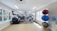 This fitness center in Diamond offers a thoughtfully designed single-floor workout space filled with a comprehensive range of exercise equipment. The room features a row of cardio machines, including ellipticals and treadmills from Life Fitness, arranged along one side to maximize floor space. Strength and resistance training equipment lines the opposite area, providing options for a full-body workout. Large windows along the walls flood the space with abundant natural light, creating an open and energizing atmosphere. The ceiling is fitted with recessed fluorescent lighting and a ceiling fan, ensuring consistent illumination and airflow throughout. The flooring combines light hardwood and patterned carpet tiles, offering both functional and aesthetic appeal. Exercise balls in varying sizes are neatly stored near the windows, adding versatility to the available workout options. The clean white walls and bright interior contribute to an inviting, motivating environment. The overall layout is spacious and organized, allowing for comfortable movement between equipment stations while maintaining an uncluttered, professional feel suited for a range of fitness routines.