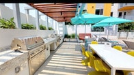 This outdoor amenity space is thoughtfully designed for communal entertaining and relaxed outdoor living. A wood-slat pergola overhead provides partial shade while allowing natural light to filter through, creating a bright and inviting atmosphere across the light-toned tile flooring. The outdoor kitchen is a standout feature, equipped with a stainless steel gas grill, granite countertops, and a utility sink — offering everything needed for convenient outdoor cooking and food preparation. Raised planter beds line the perimeter walls, filled with lush tropical greenery that adds color and a sense of privacy to the enclosed space. The dining area is furnished with long white tables and vibrant yellow chairs, complemented by teal market umbrellas that inject a playful, modern aesthetic. Wall-mounted speakers are integrated into the space, supporting an audio-ready environment for gatherings. Views from the perimeter reveal a nearby pool area and surrounding multi-story building facades, suggesting this space functions as a shared outdoor amenity within a larger residential community. Overall, the space balances functional design with lively, contemporary styling.