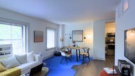 This single-floor space showcases an open-plan layout that seamlessly integrates a living area and dining space. The interior is characterized by clean white walls that enhance the sense of brightness throughout, complemented by warm hardwood flooring that anchors the space with a natural aesthetic. A striking cobalt blue area rug defines the dining zone, adding a bold pop of color that contrasts effectively with the neutral surroundings.

The dining area features a wooden table paired with a mix of modern chairs, including a classic Wishbone-style chair and contemporary molded seating, reflecting an eclectic yet cohesive design sensibility. Wall-mounted swing-arm sconces and a floor lamp provide layered ambient lighting, while framed artwork adorns the walls, contributing to the curated aesthetic.

The adjacent living area includes a white sofa and coffee table, maintaining the space's clean, minimalist character. A window with a simple blind allows natural light to filter in, brightening the space. Overall, the interior presents a modern, well-appointed environment with thoughtful design details and a welcoming ambiance.