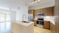 This single-floor space showcases a clean, contemporary design with an open-concept layout that seamlessly connects the kitchen and living area. The kitchen is a standout feature, equipped with warm wood-toned cabinetry extending to the ceiling for ample storage, white quartz countertops, and a full suite of stainless steel appliances including a french-door refrigerator with ice and water dispenser, an electric range, and a built-in microwave. A large central island with an undermount sink and a sleek industrial-style pull-down faucet serves as both a prep area and a casual gathering space, accented by modern pendant lighting overhead. A white subway tile backsplash adds a refined touch to the cooking zone. Light hardwood flooring flows throughout the main living areas, complementing the bright white walls and creating a warm, airy ambiance. Track lighting in the kitchen provides focused illumination, while the elevated ceilings contribute to an open, expansive feel. The bedroom offers plush carpeting, neutral tones, and generous natural light through dual windows with blinds. The bathroom features large-format tile flooring, a sleek vanity with dark cabinetry and a white countertop, and a glass-enclosed shower, maintaining the space's modern aesthetic throughout.