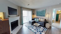 This well-appointed 2-bedroom, 2-bathroom single-floor residence offers a thoughtfully designed layout with modern touches throughout. The inviting living room serves as the heart of the home, anchored by a classic white brick fireplace that adds warmth and character. Large sliding glass doors dressed with vertical blinds and teal accent curtains flood the space with natural light while providing access to an outdoor area. Rich hardwood-style flooring flows throughout the living space, complemented by neutral wall tones that create a clean, versatile backdrop. A wall-mounted flat-screen television and tasteful décor complete the living area's polished look. The textured ceiling adds a traditional architectural element consistent throughout the home. The space is designed with both comfort and functionality in mind, offering generous room proportions that accommodate relaxed living and entertaining. The two bedrooms provide private retreats, while the dual bathrooms add everyday convenience. Overall, the residence strikes a balance between classic design elements — such as the brick fireplace — and contemporary finishes, resulting in a cohesive and welcoming living environment suited for a variety of lifestyles.