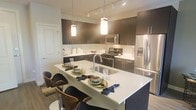 This single-floor residence showcases a thoughtfully designed open-concept layout that emphasizes both style and functionality. The kitchen serves as the centerpiece of the space, featuring rich dark cabinetry with sleek hardware that creates a striking contrast against the bright white quartz countertops and light-toned subway tile backsplash. A generous kitchen island doubles as a breakfast bar, accommodating multiple stylish modern barstools, making it an ideal spot for casual dining and entertaining. Pendant lighting hangs above the island, casting a warm, inviting glow that complements the track lighting overhead. The kitchen is outfitted with a full suite of stainless steel appliances, including a gas range, built-in microwave, and a French door refrigerator with an ice and water dispenser. The undermount sink is seamlessly integrated into the island, adding to the clean, uncluttered aesthetic. Wood-look flooring runs throughout, tying the space together with a warm, contemporary feel. The overall ambiance is modern and polished, with a careful balance of dark and light tones that lend the space an upscale, sophisticated character.