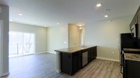 This single-floor space showcases a thoughtfully designed open-concept layout that integrates the kitchen and living areas into one cohesive environment. The kitchen serves as the focal point, anchored by a generous island with a light granite-toned countertop, an undermount sink, and dark espresso cabinetry that extends along one wall. A stainless steel dishwasher is integrated beneath the island, adding a practical, modern touch. Recessed lighting illuminates the kitchen area, complementing the flush-mount fixture visible toward the rear of the space.

The flooring throughout is a warm wood-look laminate or luxury vinyl plank, providing a durable and visually cohesive surface that ties the kitchen and living areas together. Neutral light gray walls keep the atmosphere bright and versatile. The open living area beyond the island offers generous room to furnish and personalize, with a door providing access to natural light on one side. A paneled interior door at the rear leads to an additional room or hallway, adding functional connectivity to the layout. The overall ambiance is clean, modern, and move-in ready.