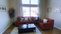 This single-story space centers around a welcoming living room designed for both comfort and functionality. The room's most prominent feature is a large picture window framed by elegant white drapes, which floods the space with abundant natural light and offers views of the surrounding neighborhood. The window's generous proportions contribute to an open, airy atmosphere throughout the room.

The living room is furnished with a bold red sofa accented by decorative floral and patterned throw pillows, complemented by a matching red accent chair. A dark espresso coffee table serves as the room's anchor, paired with a coordinating end table that holds a classic table lamp. The seating arrangement is grounded by a light-toned geometric-patterned area rug, adding texture and visual interest to the space.

The flooring appears to be a warm-toned hard surface, contributing to the room's overall sense of warmth. A large potted plant in the corner introduces a natural element, enhancing the livability of the space. The overall ambiance is cozy yet polished, with a cohesive color palette that balances rich, warm tones with lighter neutral accents.