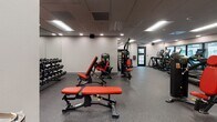 Located at 414 N Fifth Ave in Tucson, Arizona, this modern fitness center is designed to support a comprehensive range of workout routines within a single, well-organized floor. The space is finished with durable rubber flooring throughout, providing both traction and impact absorption for users across all activity zones.

The cardio area is lined with a row of treadmills and elliptical machines equipped with digital screens, positioned alongside large glass windows that bring in natural light and offer views into adjacent areas, creating an open and energizing atmosphere. A drop ceiling with recessed and panel lighting ensures consistent, bright illumination across the entire facility.

The strength training zone features a robust selection of Hoist cable machines and Matrix weight benches with red upholstery, complemented by a full rack of dumbbells along the mirrored wall — catering to both machine-based and free weight training. A standout architectural feature is the built-in climbing wall, adding a unique functional fitness element to the space.

A water fountain is conveniently installed near the entry corridor, which also provides glass-door access to adjacent building spaces. The facility maintains a clean, professional aesthetic with neutral gray and white walls, a drop-panel ceiling, and a practical, clutter-free layout designed to maximize usability and flow for all fitness levels.