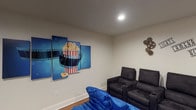 This single-floor space is thoughtfully divided into two distinct functional areas: a private movie theater and a business center. The theater room is designed with an authentic cinematic atmosphere, featuring rows of plush black recliner chairs equipped with built-in cup holders, complemented by oversized blue bean bag seating in the front for a more casual viewing experience. A large wall-mounted flat-screen television serves as the focal point, while a classic red popcorn cart adds a charming, nostalgic touch. Cinema-themed décor enhances the ambiance throughout, including a multi-panel popcorn artwork, decorative film reel wall accents, and a "Lights, Camera, Action" wall installation. Recessed ceiling lighting provides a warm, controlled glow suited for screenings. Warm-toned hardwood flooring runs throughout both areas, adding a cohesive aesthetic. The adjacent business center offers a practical workspace featuring a shared workstation table with multiple computer monitors, ergonomic mesh chairs, and a decorative wood-panel wall accent. A large window in the business center allows natural light to filter in, offering a view of the exterior. Together, both areas create a versatile, well-appointed space balancing productivity and entertainment.