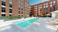 Located at 55 Romney St in Charleston, South Carolina, this outdoor amenity space is set within a modern multi-story development that blends classic red brick facades with contemporary steel-and-glass architecture. The centerpiece of the courtyard is a sleek, resort-style swimming pool with crystal-clear blue water, framed by an expansive concrete pool deck finished in large-format pavers. The surrounding sun deck is generously appointed with cushioned chaise lounges, oversized shade umbrellas, and modular outdoor seating arrangements, creating an inviting atmosphere for relaxation and socializing. Raised planter beds filled with ornamental grasses and young trees add a touch of greenery and visual texture to the hardscape. A dedicated turf area, bordered by exposed brick and positioned beneath covered balconies, provides a functional pet-friendly green space within the courtyard. The multi-story building wings enclosing the space feature floor-to-ceiling windows and private balconies, allowing natural light to fill the courtyard throughout the day while affording residents elevated views of the amenity level below. The overall design balances industrial heritage with modern outdoor living, offering a well-equipped and thoughtfully landscaped retreat in the heart of Charleston.