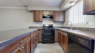 This 840-square-foot apartment features a single-floor layout with two bedrooms and two bathrooms. The open-concept living room connects directly to the kitchen, which includes a peninsula counter with dark granite-style countertops, medium-toned wood cabinetry, a gas range, and black refrigerator, with wood-style flooring underfoot. The living area features neutral carpet, crown molding, and a ceiling fan with light fixture. Both bedrooms are carpeted and equipped with ceiling fans; one includes a mirrored sliding closet, while the primary bedroom at 180 sq ft offers a walk-in closet. The primary bathroom has a wood-vanity with dark countertop and wood-style flooring. The second bathroom includes a soaking tub with tile surround. Neutral gray walls and textured ceilings are consistent throughout.