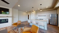 Located at 4604 Hammersley Rd in Madison, Wisconsin, this single-floor communal space offers a well-designed, open-concept layout that seamlessly integrates a kitchen, lounge, and dining areas. The interior is finished with warm wood-look flooring throughout, complemented by neutral-toned walls that create a clean, contemporary atmosphere.

The kitchen is a standout feature, equipped with stainless steel appliances, a microwave, and a generous granite-topped island with an undermount sink. White subway tile provides a striking backsplash, while white cabinetry and bar-height seating at the island make the space both functional and inviting. Pendant lighting adds a modern touch above the island.

The lounge area is furnished with a comfortable sofa, accent chairs in warm amber tones, and a coffee table set atop a patterned area rug. A white brick fireplace with a floating wood mantel serves as the focal point of the room, with a wall-mounted television above. Ceiling fans provide comfortable airflow throughout.

Large windows flood the space with natural light and offer views of the surrounding landscape, while a sliding glass door opens directly onto an outdoor balcony, extending the usable space and enhancing the overall sense of openness. A dining table with seating rounds out this versatile, well-appointed gathering space.