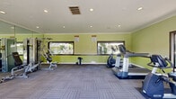 Located at 727 S Lyon St in Santa Ana, California, this fitness center offers a well-equipped workout space designed to meet a range of exercise needs. The room is finished with vibrant chartreuse green walls accented by a dark wainscoting border, crown molding, and a white ceiling with recessed lighting that keeps the space bright and energizing. Patterned carpet tiles cover the floor throughout, providing a comfortable and durable surface for various workout activities. A full-length wall mirror on one side visually expands the room and aids in proper exercise form. Large horizontal windows along two walls, along with a set of dark-framed French doors, flood the interior with abundant natural light and offer open views of the landscaped outdoor grounds and pool area beyond. The fitness center is equipped with multiple cardio machines, including a treadmill, elliptical trainers, and a stationary cycle, as well as a multi-station strength training unit and free weights. A punching bag and additional accessories round out the available equipment. The open layout allows for comfortable movement between stations, making this a functional and inviting facility for residents seeking convenient access to fitness amenities in Santa Ana.