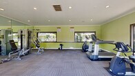 Located at 727 S Lyon St in Santa Ana, California, this fitness center offers a well-equipped workout space designed to meet a range of exercise needs. The room is finished with vibrant chartreuse green walls accented by a dark wainscoting border, crown molding, and a white ceiling with recessed lighting that keeps the space bright and energizing. Patterned carpet tiles cover the floor throughout, providing a comfortable and durable surface for various workout activities. A full-length wall mirror on one side visually expands the room and aids in proper exercise form. Large horizontal windows along two walls, along with a set of dark-framed French doors, flood the interior with abundant natural light and offer open views of the landscaped outdoor grounds and pool area beyond. The fitness center is equipped with multiple cardio machines, including a treadmill, elliptical trainers, and a stationary cycle, as well as a multi-station strength training unit and free weights. A punching bag and additional accessories round out the available equipment. The open layout allows for comfortable movement between stations, making this a functional and inviting facility for residents seeking convenient access to fitness amenities in Santa Ana.
