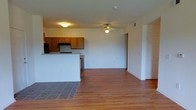 This single-floor residence offers a practical and comfortable open-concept layout that seamlessly connects the kitchen and living areas. The kitchen is equipped with warm honey-toned wood cabinetry, a black refrigerator, and a range, all complemented by a breakfast bar peninsula that provides both additional prep space and a natural dividing point between the two zones. Grey tile flooring anchors the kitchen area, creating a clear visual distinction from the rest of the space. The living area extends generously from the kitchen, featuring smooth hardwood flooring that adds warmth and continuity throughout. A ceiling fan with a light fixture is centrally mounted, providing both airflow and ambient lighting to the open room. The walls are finished in clean, neutral tones, keeping the space bright and versatile. A doorway visible to the side suggests access to additional rooms or storage. The overall layout feels open and airy, with a simple, functional design that makes efficient use of the available footprint, ideal for comfortable everyday living.
