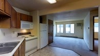 This single-floor unit offers a functional and straightforward layout, with the kitchen and living area flowing seamlessly into one another. The kitchen is equipped with essential appliances, including a white electric range, a built-in microwave, and a full-sized refrigerator. Dark wood cabinetry overhead provides ample storage, while the light-toned vinyl flooring adds a practical, easy-to-clean surface underfoot. A textured ceiling runs throughout the space, lending a classic character to the interior.

Adjacent to the kitchen, the living area transitions to wall-to-wall carpeting, creating a comfortable and defined space. Large windows along the far wall allow natural light to fill the room, contributing to an open and airy feel. A wall-mounted air conditioning unit is visible beneath the windows, complemented by baseboard heating along the perimeter for year-round climate control. A door off the main corridor leads to an additional room, offering further functional space within the unit. Overall, the layout prioritizes practicality while maintaining a comfortable and livable atmosphere.