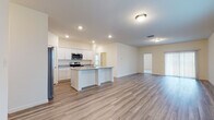 This single-story home showcases a thoughtfully designed open-concept layout that seamlessly connects the kitchen, dining, and living areas. Light-toned wood-look flooring flows throughout the main living spaces, complementing the bright, neutral color palette and recessed lighting overhead.

The kitchen serves as a central hub, anchored by a large island with granite countertops and an undermount sink. White shaker-style cabinetry lines the walls, providing ample storage, while a full suite of stainless steel appliances — including a French door refrigerator, electric range, built-in microwave, and dishwasher — complete the space.

The expansive living room receives generous natural light through multiple windows and a sliding glass door, creating an airy, open atmosphere. Bedrooms feature plush carpeting and standard closets with hanging rods, offering practical storage solutions. One bedroom benefits from direct access to a bathroom equipped with a walk-in shower and wood-look tile flooring.

A dedicated laundry room includes overhead shelving, washer and dryer hookups, and a window, adding both functionality and natural light. Throughout, the home maintains a clean, modern aesthetic with consistent finishes and a bright, welcoming ambiance.