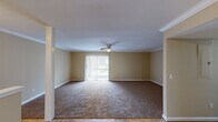 This single-floor residence offers a thoughtfully laid-out interior with a warm, neutral color palette carried consistently throughout. The living room is generously sized, featuring plush wall-to-wall carpeting, crown molding, and a ceiling fan with integrated lighting. A sliding glass door floods the space with natural light and provides direct access to an outdoor area, complemented by vertical blinds for privacy and light control. Adjacent to the living room, the dining area transitions seamlessly via warm-toned tile flooring, accented by elegant crown molding and a flush-mount ceiling light. An open pass-through connects directly to the kitchen, which is equipped with rich wood cabinetry, dark granite countertops, a stainless steel double-basin sink, a dishwasher, and an electric range — all anchored by a window that invites natural light. The bedrooms are carpeted and comfortably proportioned, each featuring windows with blinds that provide views of the surrounding exterior. The layout, as visible in the dollhouse view, is compact yet efficiently organized, offering clear spatial flow between the living, dining, kitchen, and sleeping areas. The overall ambiance is clean, functional, and move-in ready.