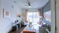 This single-floor space offers a thoughtfully designed living area that balances comfort and modern style. The living room serves as the centerpiece, featuring dark hardwood flooring that contrasts beautifully with the light gray walls, creating a clean and contemporary aesthetic. A ceiling fan with an integrated light fixture is centrally positioned overhead, providing both airflow and ambient lighting to the space.

Natural light pours in through a large sliding glass door flanked by warm, patterned orange and white curtains, offering views of the outdoors and lending the room an airy, open feel. The layout accommodates a generous seating arrangement, including a plush sectional sofa accented with patterned throw pillows in complementary orange and gray tones, a coordinating accent chair, and a sleek round coffee table set.

A console table along one wall provides additional surface space and decorative display opportunities, styled with books, accent pieces, and a striking floral arrangement. Wall-mounted shelving and curated artwork add character and depth to the room. The overall ambiance is warm and inviting, with a cohesive color palette of white, gray, and orange that gives the space a polished, move-in-ready feel throughout.