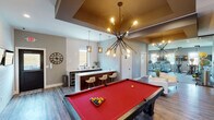 This thoughtfully designed single-floor amenity space blends recreation, relaxation, and fitness into one cohesive environment. The central billiards lounge serves as the heart of the space, anchored by a full-size pool table beneath a striking sputnik-style chandelier. A coffered ceiling detail with a warm gold finish adds architectural depth and defines the gaming area. Shiplap accent walls and dual wall-mounted flat-screen TVs enhance the entertainment-focused atmosphere. Rich wood-look plank flooring runs throughout, tying each zone together seamlessly. Adjacent to the billiards area, a sleek wet bar features a stainless steel sink, quartz countertops, dark wood cabinetry, and bar seating beneath pendant lighting, providing a convenient refreshment station. A dramatic reclaimed wood accent wall houses both a large flat-screen TV and a linear electric fireplace below, creating a warm and inviting lounge nook furnished with contemporary white armchairs. Glass partition walls offer a transparent boundary to the well-equipped fitness center, which features cardio machines, free weights, and mirror-lined walls. Natural light filters in through windows, and the overall ambiance strikes a balance between casual comfort and polished, modern design throughout.