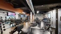 Located at 200 McFarland St in Cincinnati, Ohio, this fitness center offers a comprehensive workout environment designed to accommodate a wide range of training styles. The space features an industrial aesthetic, with exposed ductwork running along a dark ceiling, painted brick walls in white and warm orange accents, and durable rubber flooring throughout.

The strength training area is generously appointed with a full rack of dumbbells, multiple benches, cable machines, a multi-functional power rack, and a barbell station — providing ample options for both free weight and machine-based workouts. Large wall mirrors line portions of the gym, enhancing the sense of space and allowing members to monitor form during exercises.

A dedicated cardio zone includes treadmills, stair climbers, and rowing machines, positioned near large frosted windows that allow natural light to filter in. A standout feature of the space is a colorful climbing wall, adding a dynamic and functional training element not commonly found in standard fitness facilities.

Ceiling fans and mounted flat-screen televisions are distributed throughout the floor, contributing to both comfort and an engaging workout atmosphere. The overall layout is open and well-organized, making efficient use of the available floor space while maintaining clear circulation paths between equipment zones.