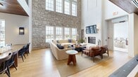 Located at 3700 Limestone Parkway in Gainesville, Georgia, this thoughtfully designed community space delivers an impressive blend of architectural drama and functional amenities. The centerpiece is a grand living room anchored by a soaring two-story stone accent wall, a fireplace, and an expansive bank of windows that flood the space with natural light and offer views of the surrounding community. Light hardwood flooring runs throughout, unifying the open layout with warmth and continuity. The living area transitions seamlessly into a spacious dining zone, where pendant lighting descends from a rich wood-clad ceiling, complementing the casual yet refined atmosphere. A well-appointed kitchen features an extended white quartz island, white cabinetry with brass hardware, and oversized globe pendant lights, all framed by additional windows overlooking the outdoor amenities. A dedicated game room offers both a billiards table and a ping-pong table beneath a striking reclaimed wood ceiling, with a staircase leading to an upper mezzanine level. A separate, intimate game niche with a tray ceiling provides a quieter setting for table games. Together, these spaces create a dynamic, welcoming environment suited for both social gatherings and relaxed everyday use.