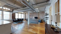 Unit 407 is a striking two-bedroom, two-bathroom loft that masterfully blends industrial character with residential comfort. The open-plan layout is immediately commanding, featuring soaring ceilings with exposed structural beams and large silver ductwork that serve as defining architectural elements. Original hardwood floors in warm honey tones flow seamlessly throughout the space, adding texture and warmth to the industrial aesthetic.

The expansive main living area is anchored by exposed brick walls, lending the unit a rich, historic charm. Large windows along the perimeter allow abundant natural light to fill the space, highlighting the interplay between the raw materials and the open floor plan. The living zone comfortably accommodates a full sectional seating arrangement, while the adjacent dining area features ample space for a large dining table ideal for entertaining.

Industrial-style open shelving lines the brick walls, offering both storage and display opportunities while reinforcing the loft's aesthetic. Structural columns are tastefully integrated into the layout, defining zones without interrupting the sense of openness. The overall ambiance is one of urban sophistication — a thoughtfully designed space that honors its architectural heritage while offering modern, comfortable living.
