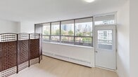 This efficiently designed one-bedroom, one-bathroom unit in California offers a bright and modern living experience across a single floor. The spacious primary bedroom is the heart of the unit, featuring light hardwood flooring, crisp white walls, and an impressive wall of floor-to-ceiling windows that frame panoramic city skyline views. A sliding door provides access to a balcony, allowing natural light to pour in throughout the day. Two side-by-side closets with bifold doors offer generous built-in storage, keeping the room clean and functional. The open layout is further enhanced by a decorative woven room divider that adds a warm, textural contrast to the otherwise minimalist aesthetic. A hallway connects the main living area to the bathroom, which is styled with a sleek, contemporary design. White beveled subway tiles line the walls, complemented by dark charcoal floor tiles and a floating vanity with a dark wood finish. A large black-framed mirror spans the width of the vanity, amplifying the sense of space, while recessed lighting adds a polished touch. The kitchen area, visible from the entry, is compact yet functional. Throughout the unit, the consistent use of light tones and clean lines creates a cohesive, modern ambiance ideal for urban living.