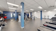 Located at 6649 Ridge Avenue in Philadelphia's Roxborough neighborhood, this fitness facility offers a generously sized, single-floor exercise room designed to support a comprehensive workout experience. The space features a clean, modern aesthetic defined by crisp white walls accented with bold blue columns and accent walls, durable speckled rubber flooring, and a suspended drop-tile ceiling with recessed fluorescent lighting that keeps the environment bright and energizing.

The exercise room is thoughtfully organized into distinct functional zones. The cardio area is lined with treadmills, elliptical machines, and stationary bikes positioned near frosted windows that allow diffused natural light to fill the space. The strength training section houses a robust selection of Hoist and other cable and weight machines, along with free weights, adjustable benches, medicine balls, and wall-mounted mirrors that enhance the sense of openness. Additional amenities include a built-in water fountain, hand sanitizer dispensers, and stainless steel waste receptacles for user convenience.

Multiple access points, including double glass entry doors, ensure smooth flow throughout the space. The open floor plan and bold color scheme create a motivating atmosphere well-suited for individual or group fitness use.