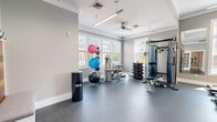 Located at 11 Kensington Ln in Rocky Hill, Connecticut, this fitness center offers a thoughtfully designed workout space that balances functionality with a clean, modern aesthetic. The single-floor layout provides a comfortable and open exercise environment, accommodating a diverse range of fitness activities without feeling crowded.

The interior features neutral gray walls complemented by warm wood plank accent walls, creating an inviting atmosphere. Durable rubber flooring covers the entire space, providing safety and support during workouts. Multiple large windows line the walls, flooding the room with natural light and offering views of the surrounding neighborhood.

Overhead, ceiling fans and a combination of pendant and linear lighting fixtures ensure consistent airflow and visibility throughout the space. A wall-mounted television is positioned near the cardio area for entertainment during workouts.

The fitness center is well-stocked with a comprehensive selection of equipment. Cardio offerings include treadmills, stationary bikes, and an elliptical machine. Strength training is supported by a multi-function cable machine, a dumbbell rack with a full range of weights, a workout bench, and various stability and medicine balls. A hydration station and a built-in bench with storage near the entrance add practical convenience. French doors lead to an adjacent area, enhancing accessibility and the overall flow of the space.