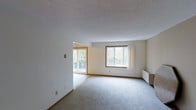 This single-floor space offers a clean, open layout filled with abundant natural light. The main living area features plush carpeting, white walls, and a textured ceiling that lend a simple, understated aesthetic. A large picture window with warm wood trim frames lush, tree-lined views, bringing the outdoors in and creating a serene atmosphere throughout the day. Adjacent to the living area, a transition to tile flooring marks a secondary space, which opens directly onto an outdoor balcony through a sliding glass door — ideal for enjoying the surrounding greenery. A baseboard heating unit positioned along the exterior wall ensures year-round comfort. The open flow between the interior zones creates a sense of spaciousness, while the neutral color palette throughout provides a versatile backdrop suitable for a variety of furnishing styles. The overall ambiance is calm and airy, with the natural wooded views serving as a defining feature of the space.